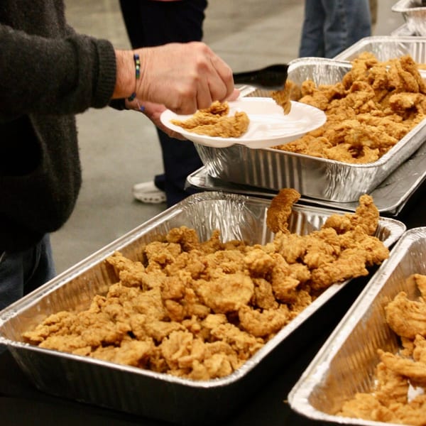 A person serves themselves crispy fried chicken tenders from large aluminum trays.