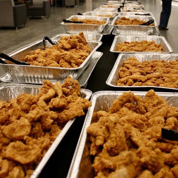 Rows of aluminum catering trays filled with golden-brown fried chicken tenders on a dark table.